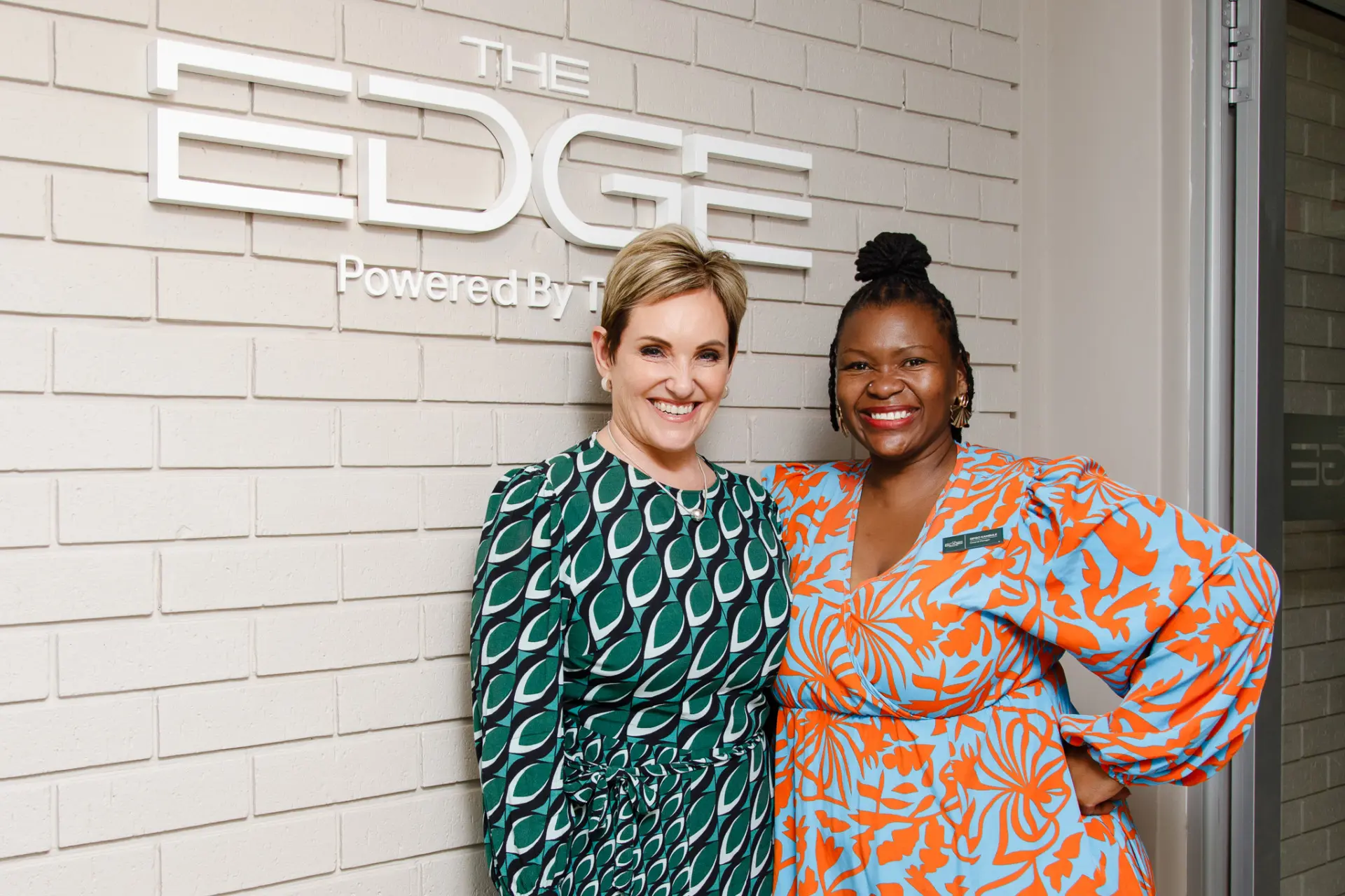 Two professional women smiling at The Edge event, one wearing green patterned dress, one in orange floral blazer.