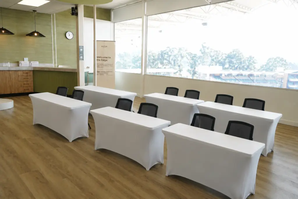 Modern conference room with three white-draped tables, black chairs, and large windows overlooking trees and stadium.