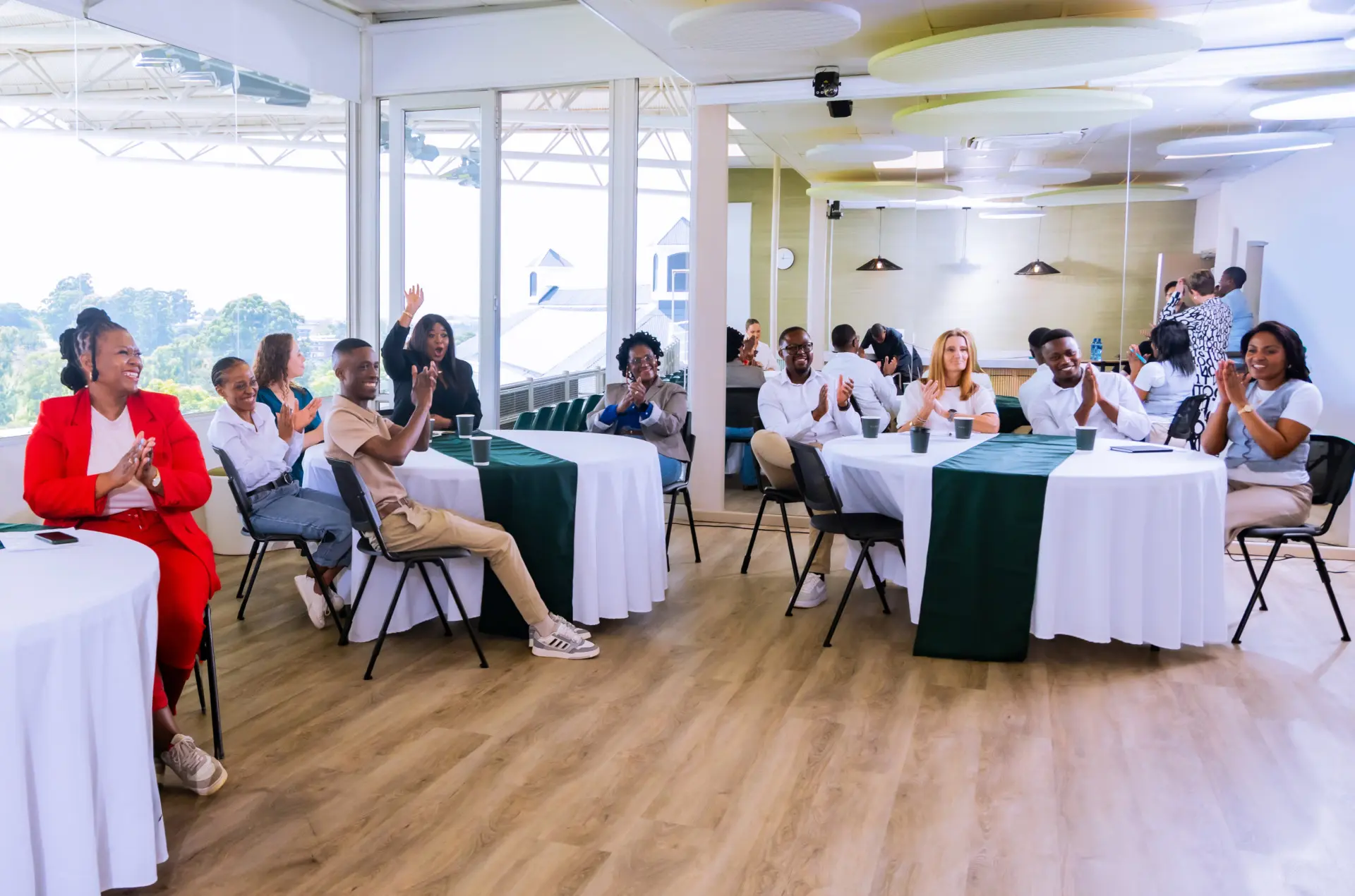 Professional diverse team applauding at modern office conference event with green tables and panoramic windows.