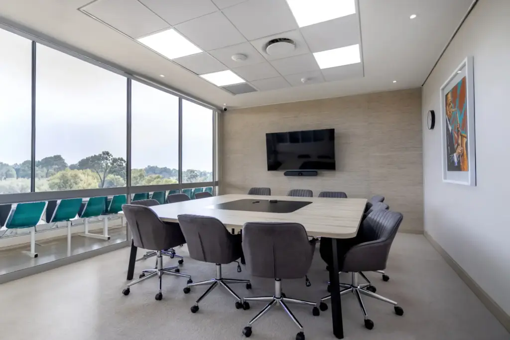 Modern conference room with oval white table, gray chairs, wall-mounted TV, and panoramic windows overlooking landscape.