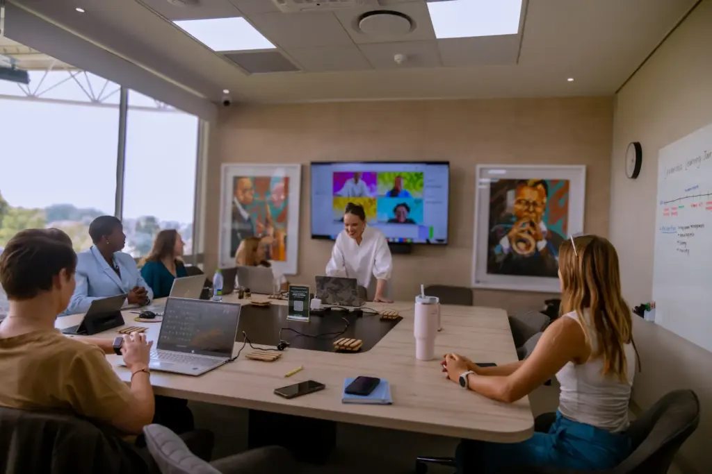 Team in modern conference room with video call on screen and laptops, colorful artwork on walls, natural light from windows.