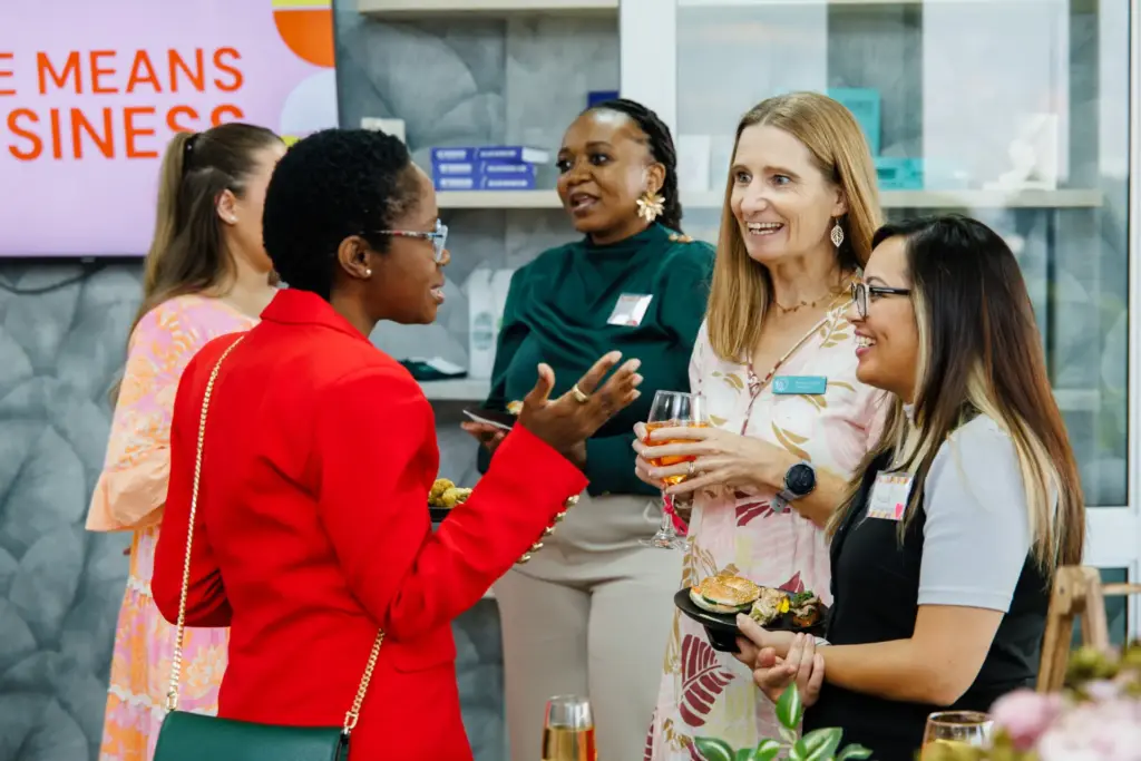 Five professional women networking at a business event, holding drinks and appetizers, smiling and conversing in a modern office setting.