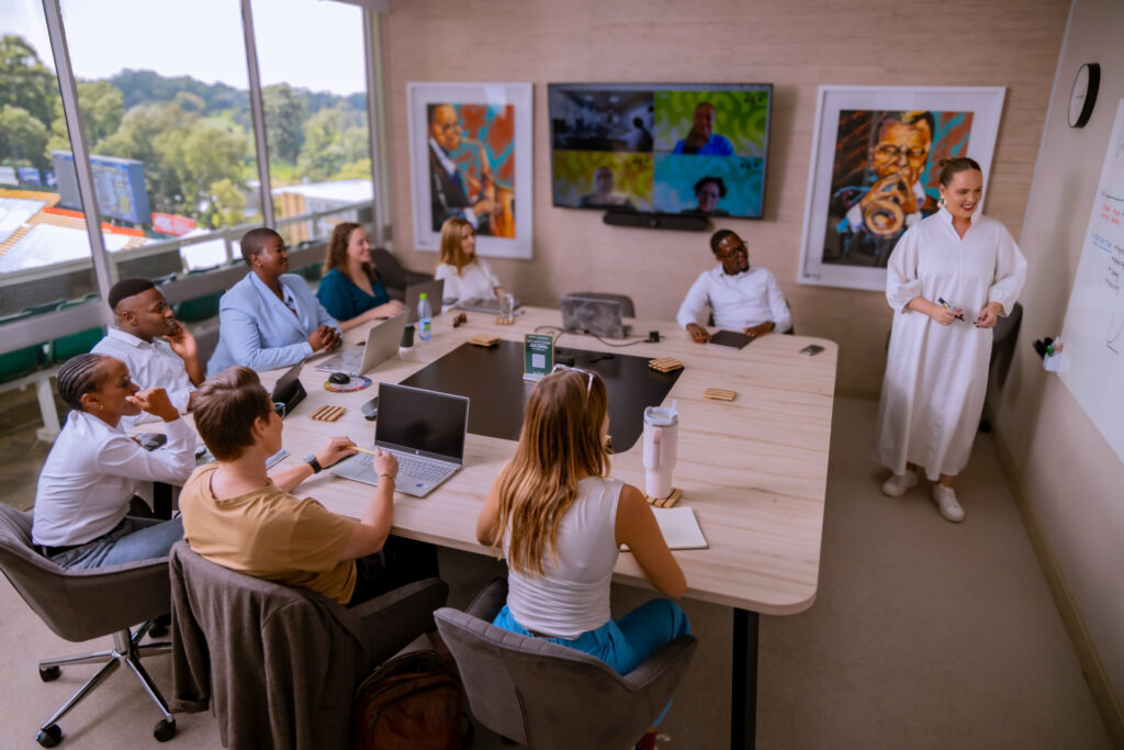 Professional team meeting in modern office with diverse group discussing around wooden table with laptops and notebooks.
