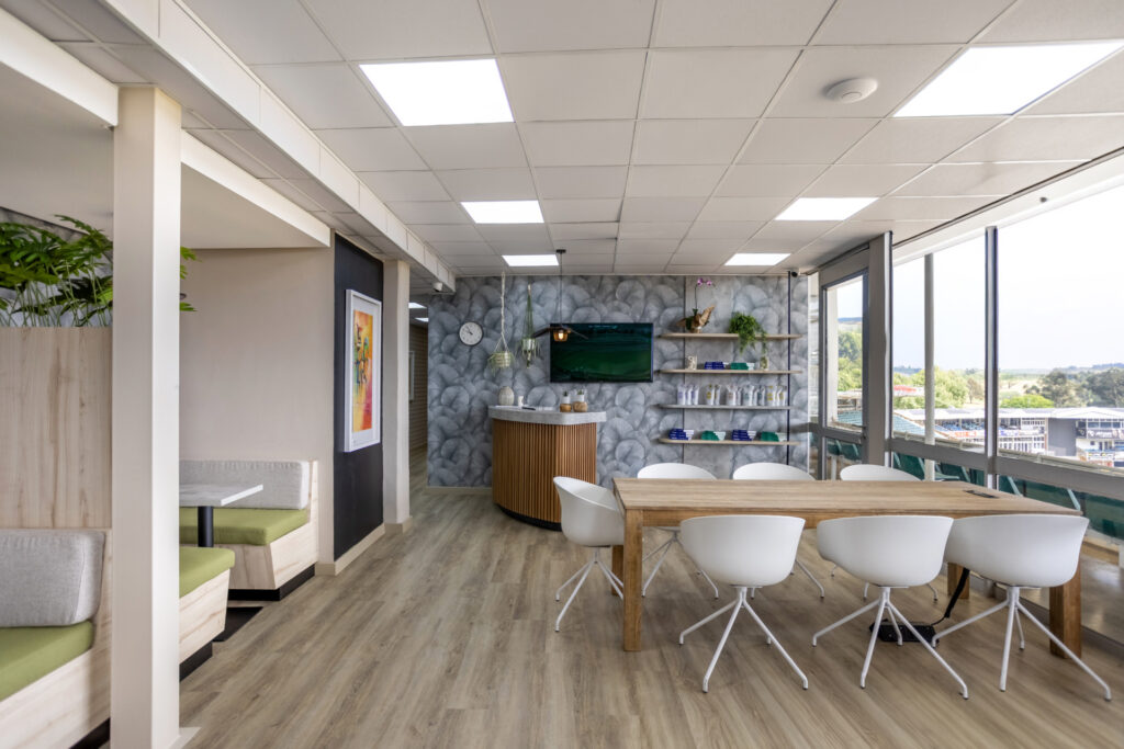 Modern office break room with wooden table, white chairs, kitchenette, and panoramic windows overlooking landscape.